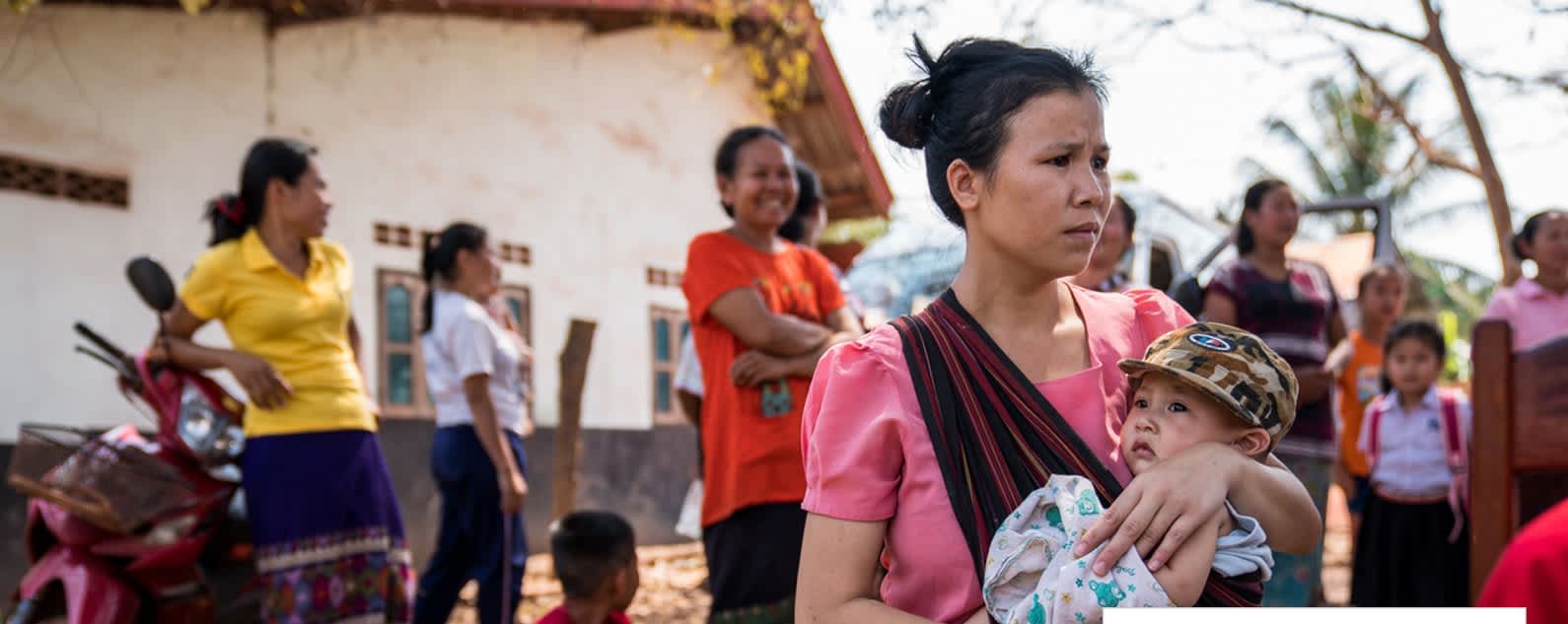 Woman carrying child in rural area. Multiple people at the background.