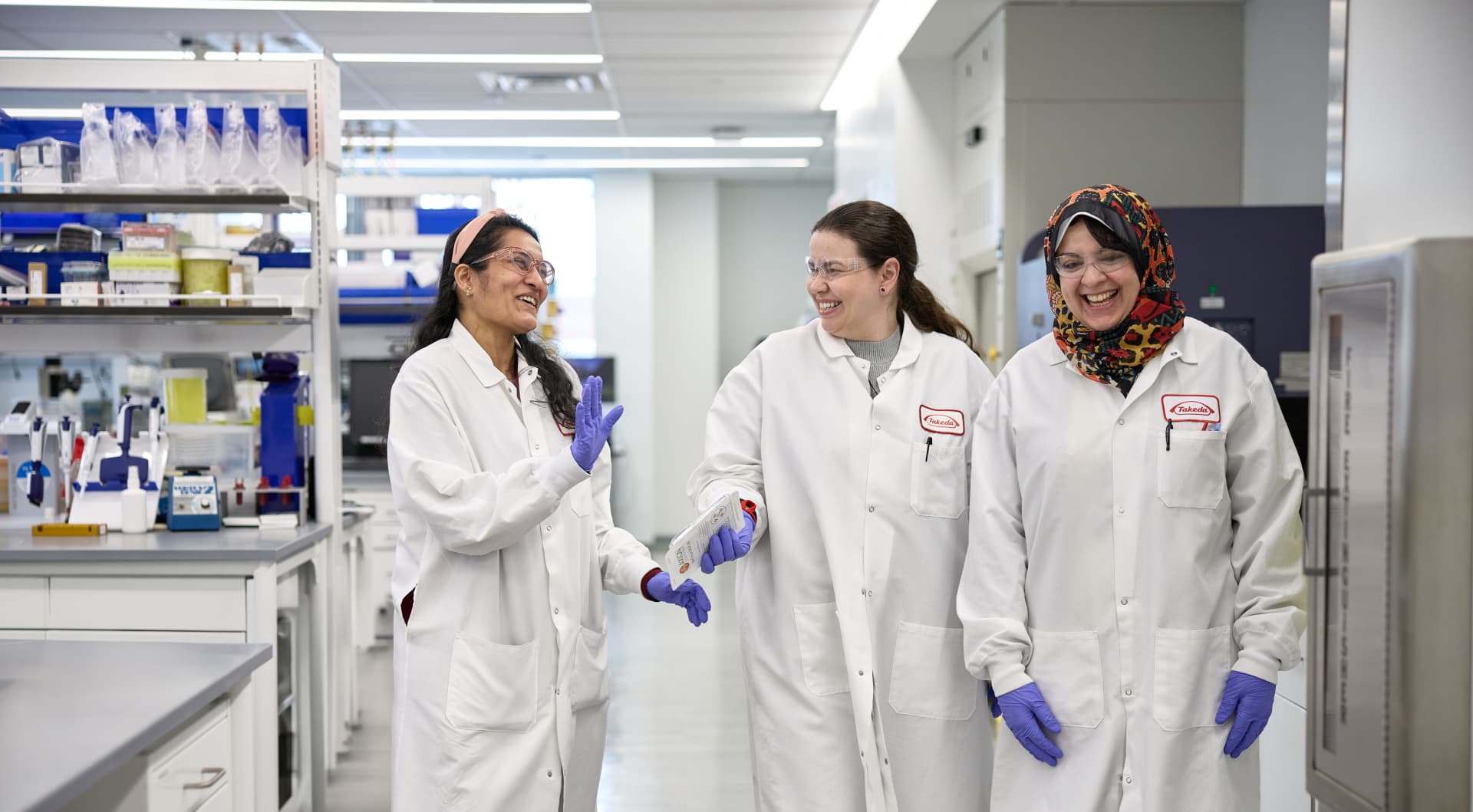 白衣を着た3人の研究者が、研究室内を歩きながら話している様子