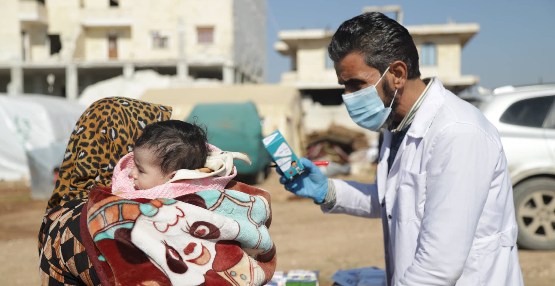 A person in a white coat holding a blue box and speaking to another person holding a child