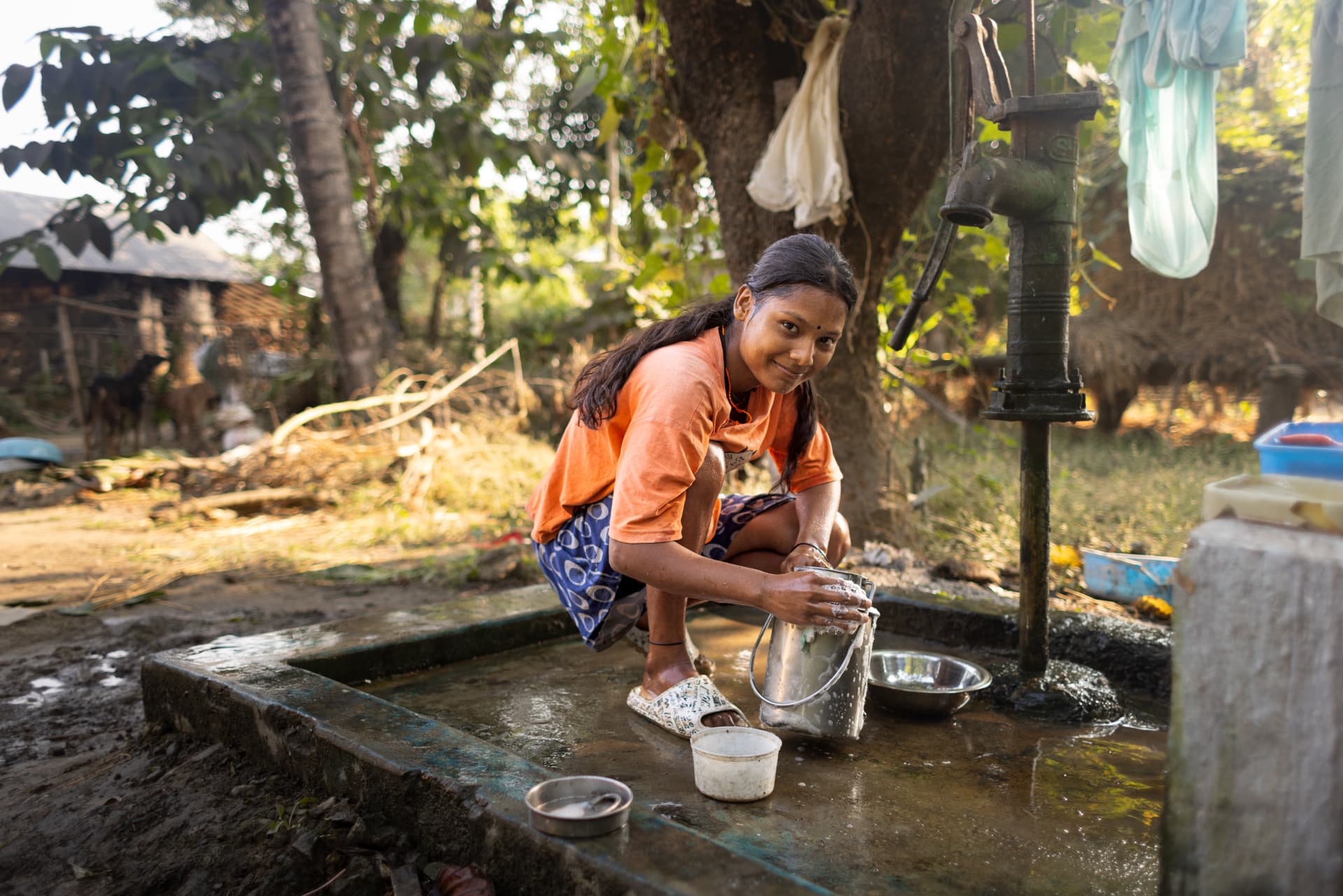 Person washing a container at a hand pump outdoors.