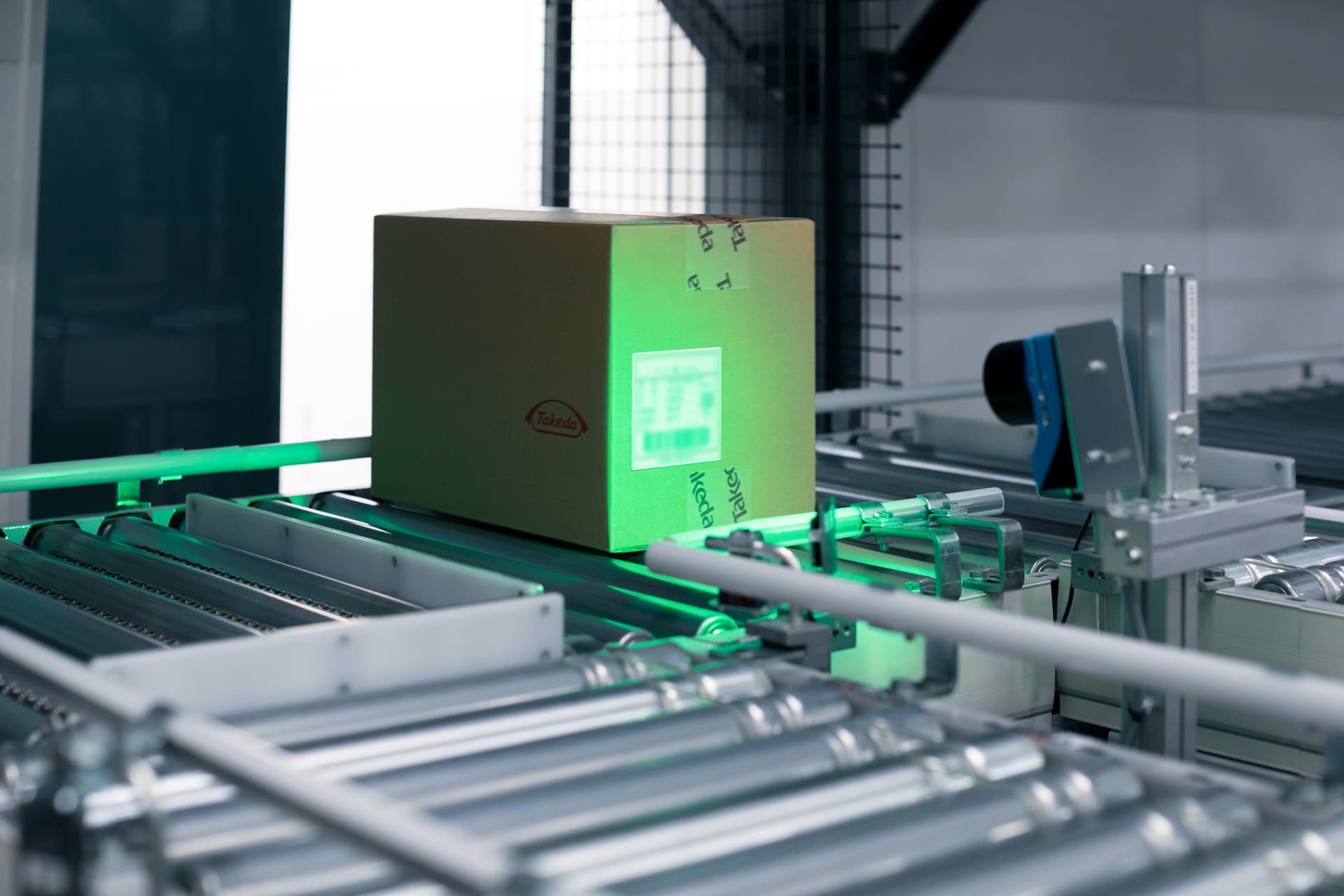 A cardboard box with a Takeda label moves along a conveyor as a scanner projects a green light to read its barcode in an automated packaging facility.