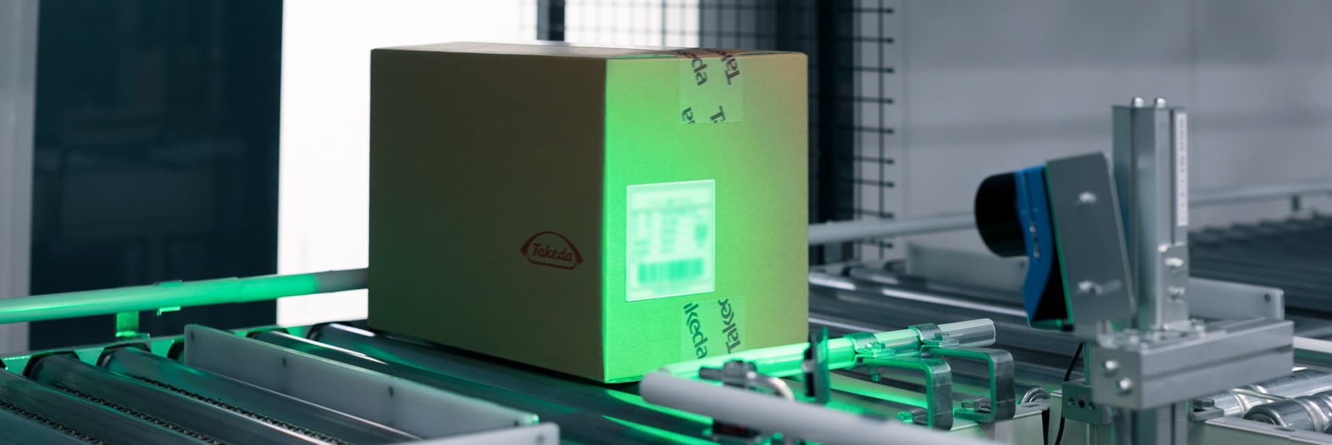 A cardboard box with a Takeda label moves along a conveyor as a scanner projects a green light to read its barcode in an automated packaging facility.