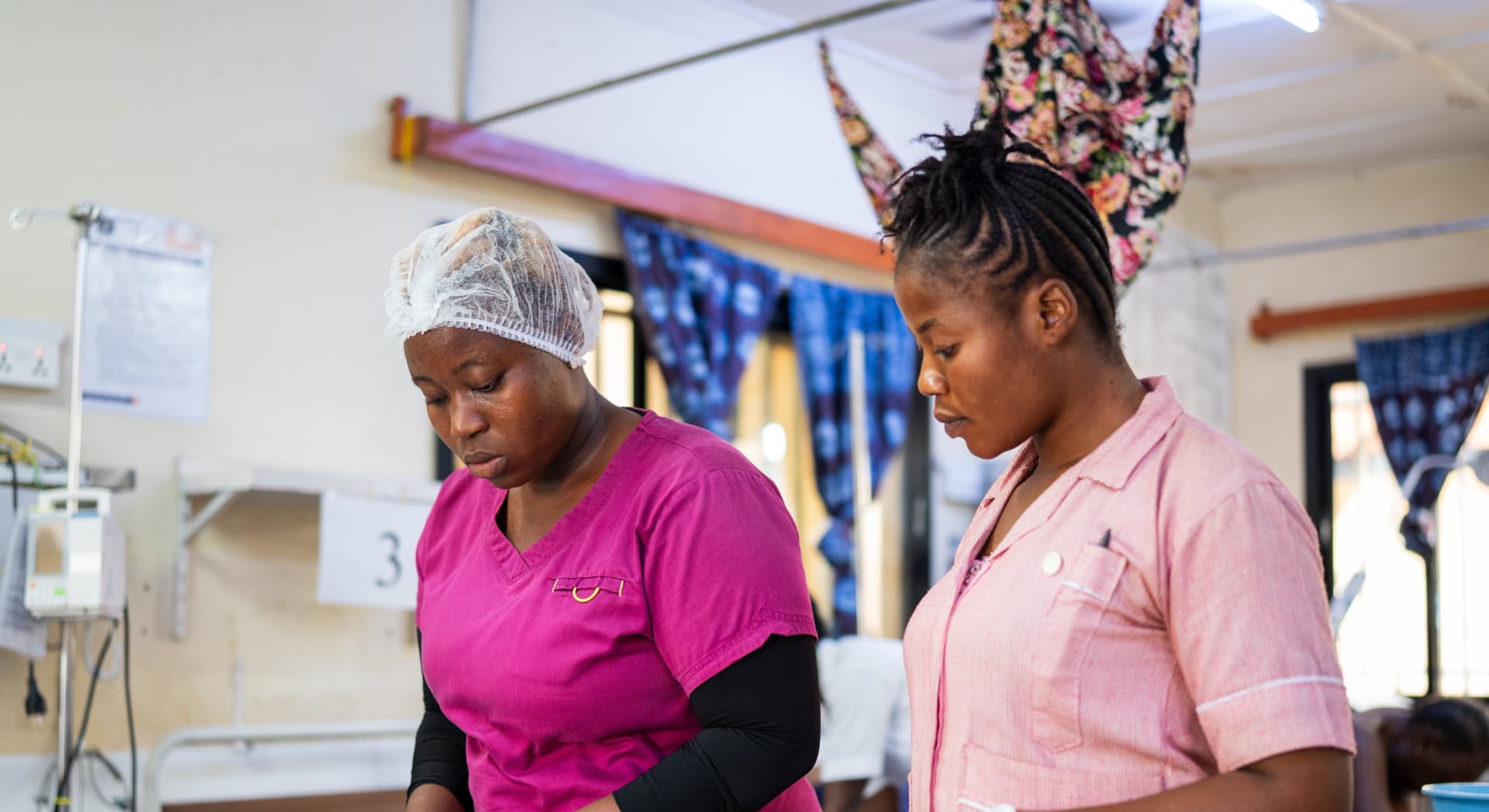 Two nurses in pink uniforms provide care to a patient in a hospital ward with medical equipment nearby.