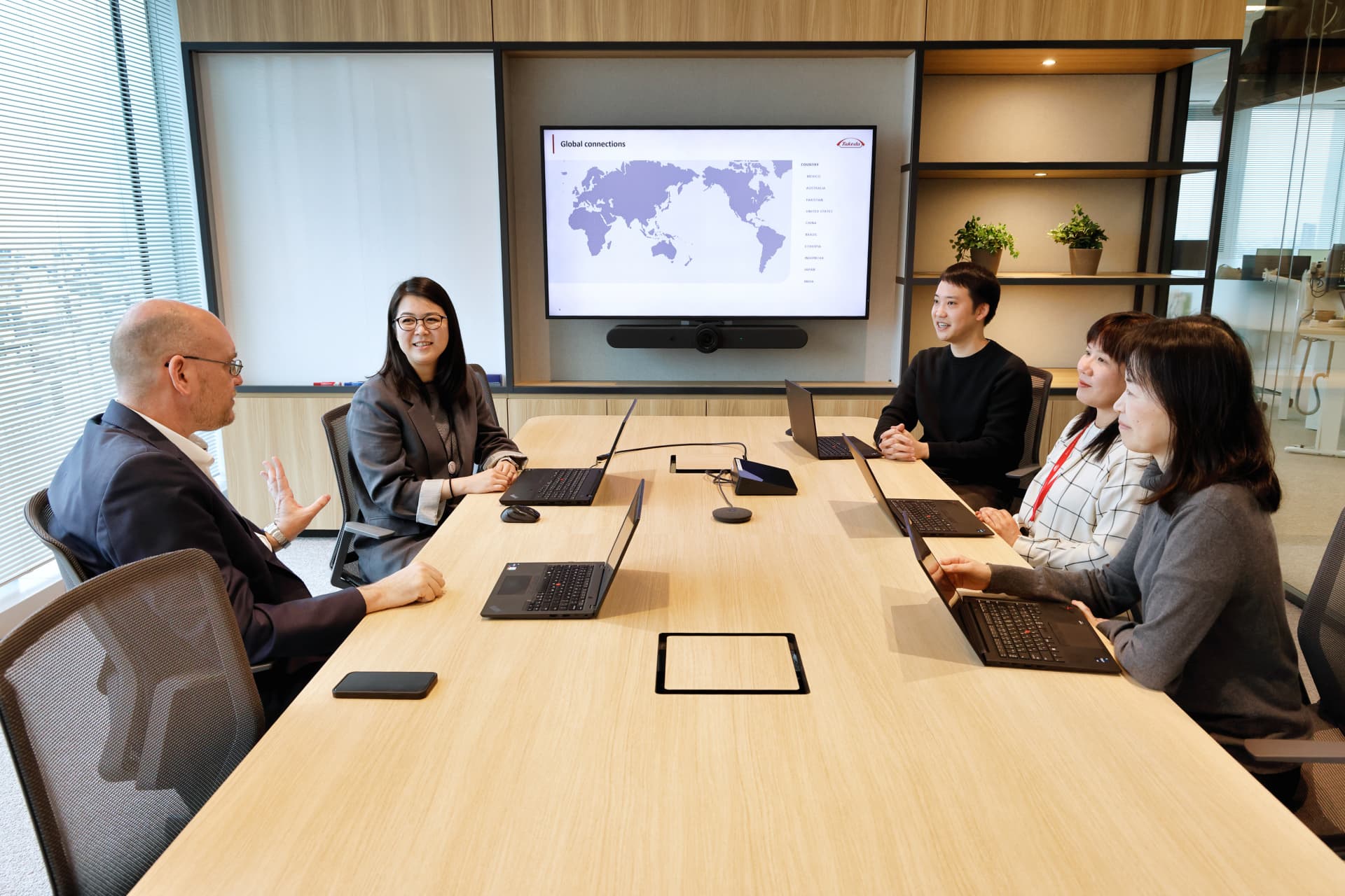 会議室で5人のビジネスパーソンがノートパソコンを使い、スクリーンを背に会議をしている様子。