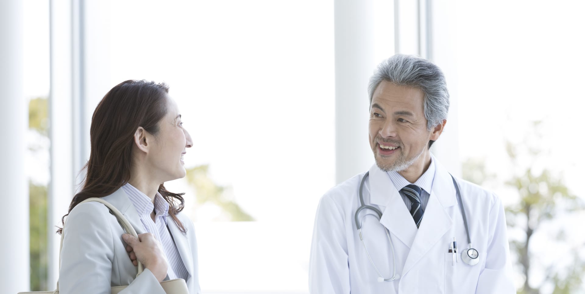 明るい屋内で、白衣を着た医療従事者がスーツ姿の来訪者と立ち話をしている様子。背景には大きな窓から自然光が差し込んでいる。