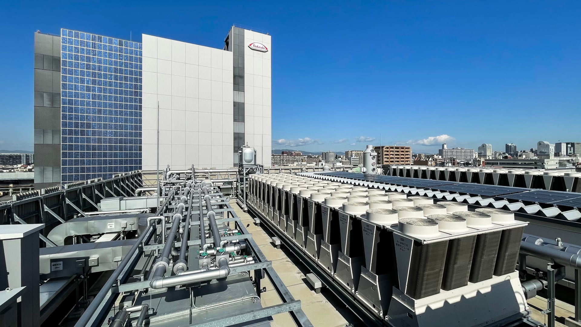 製薬工場の屋上に設置された大型空調設備と配管システム。背景にはガラス張りの高層ビルと青空が広がり、都市の建物群が見える。