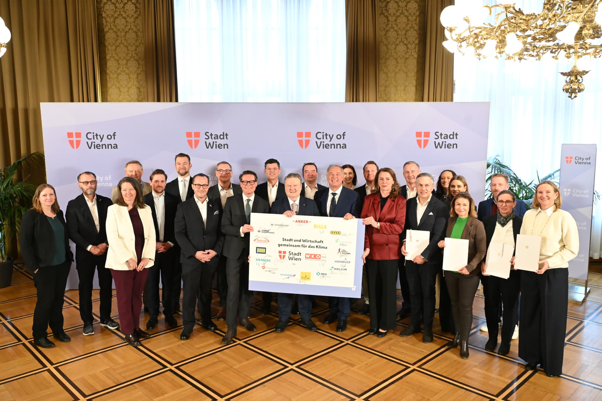 Photograph of a group of 21 people standing in a formal room holding a large check and certificates