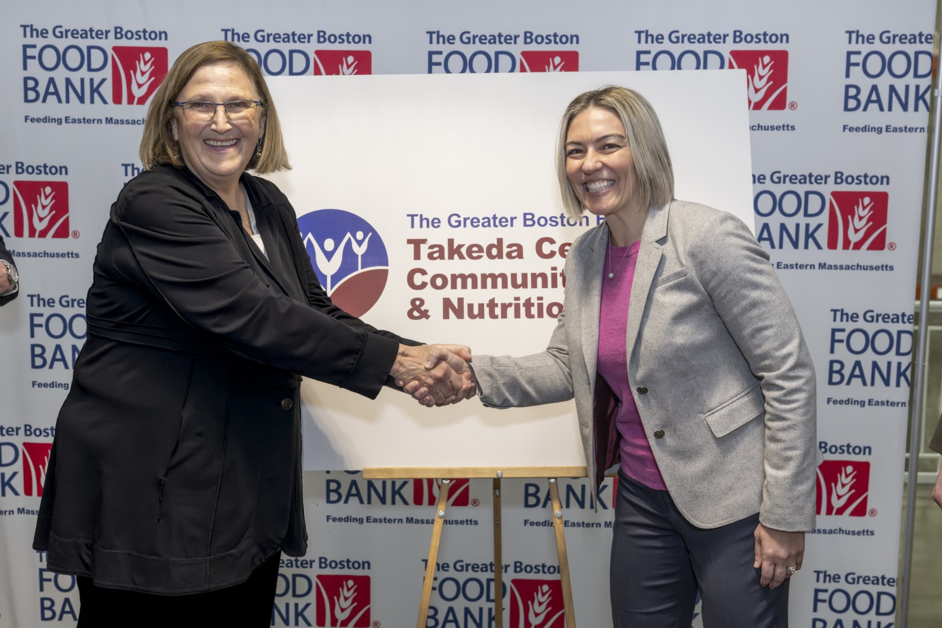 two women shaking hands in front of a sign for The Greater Boston Takeda Center for Community & Nutrition