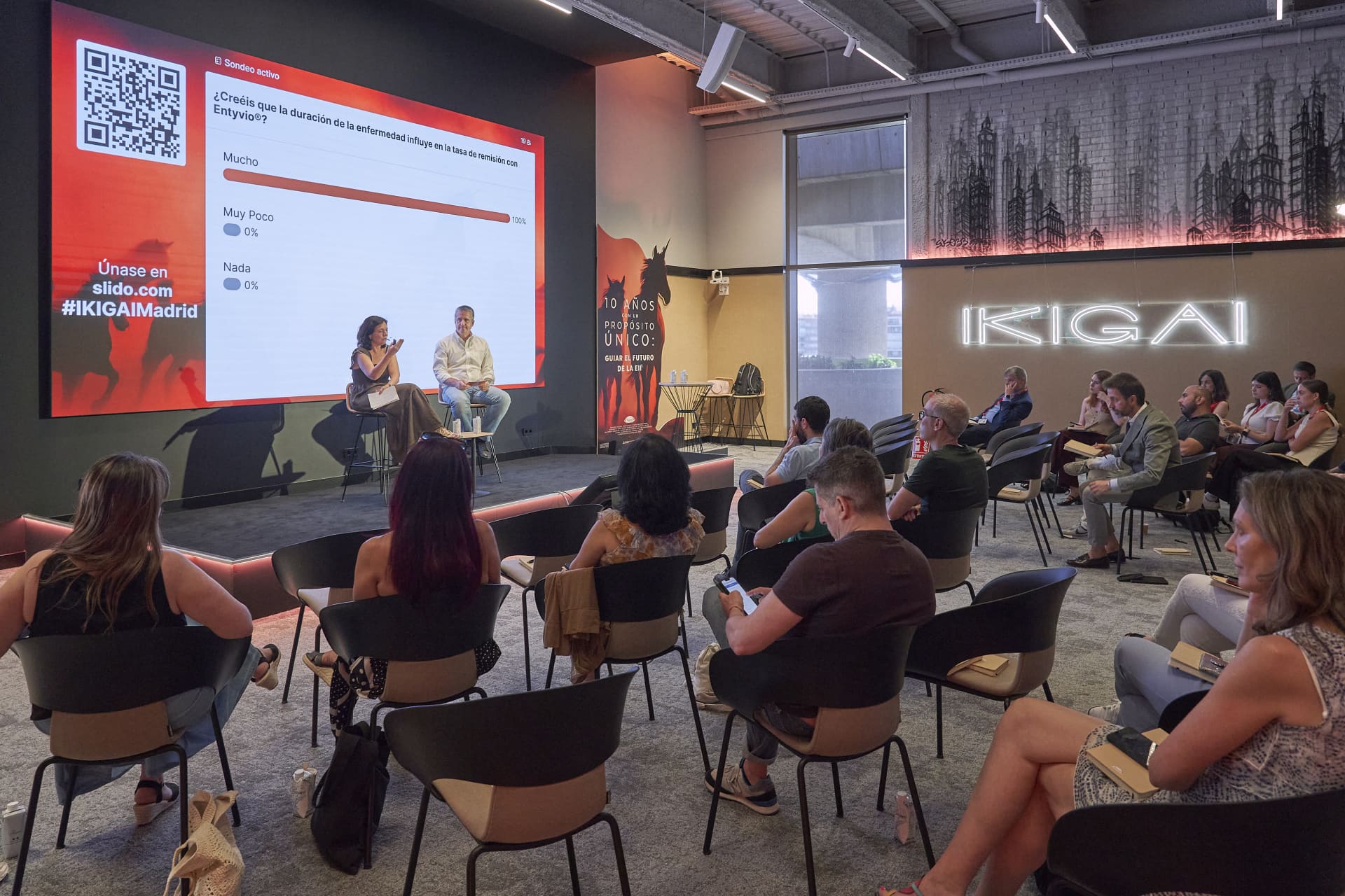 Photograph of a conference or seminar setting with attendees seated and two speakers on stage discussing a presentation slide