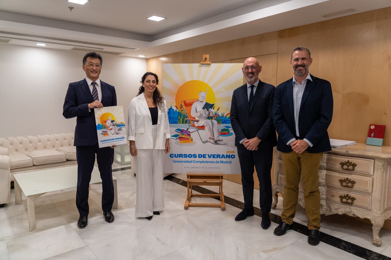 Photograph of four professionally dressed individuals standing in a room, posing next to a colorful poster on an easel promoting "Cursos de Verano Universidad Complutense de Madrid."
