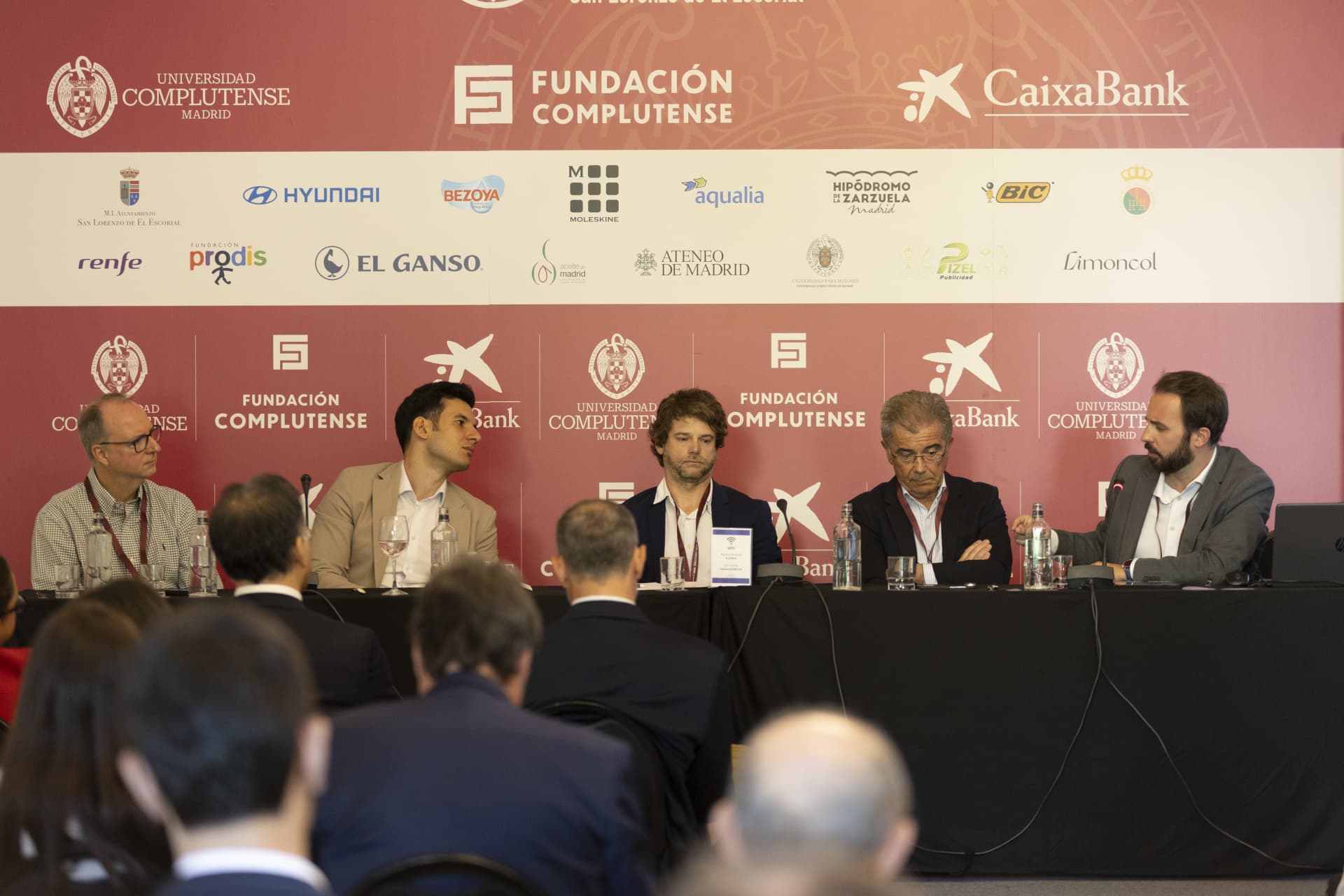 Photograph of a panel discussion at a conference featuring five men seated behind a table with microphones, with an audience in the foreground.