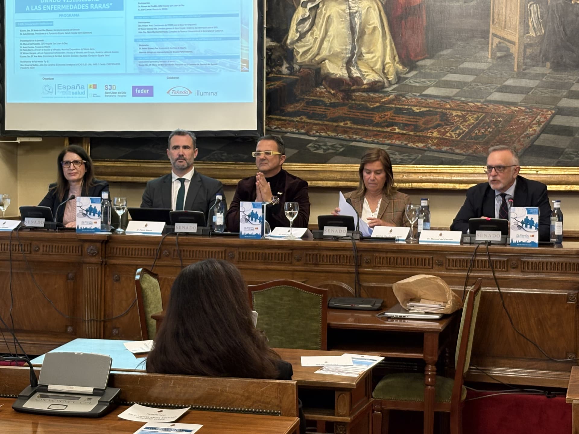 a formal panel discussion with five individuals seated behind a wooden table, each with nameplates, microphones, and informational booklet