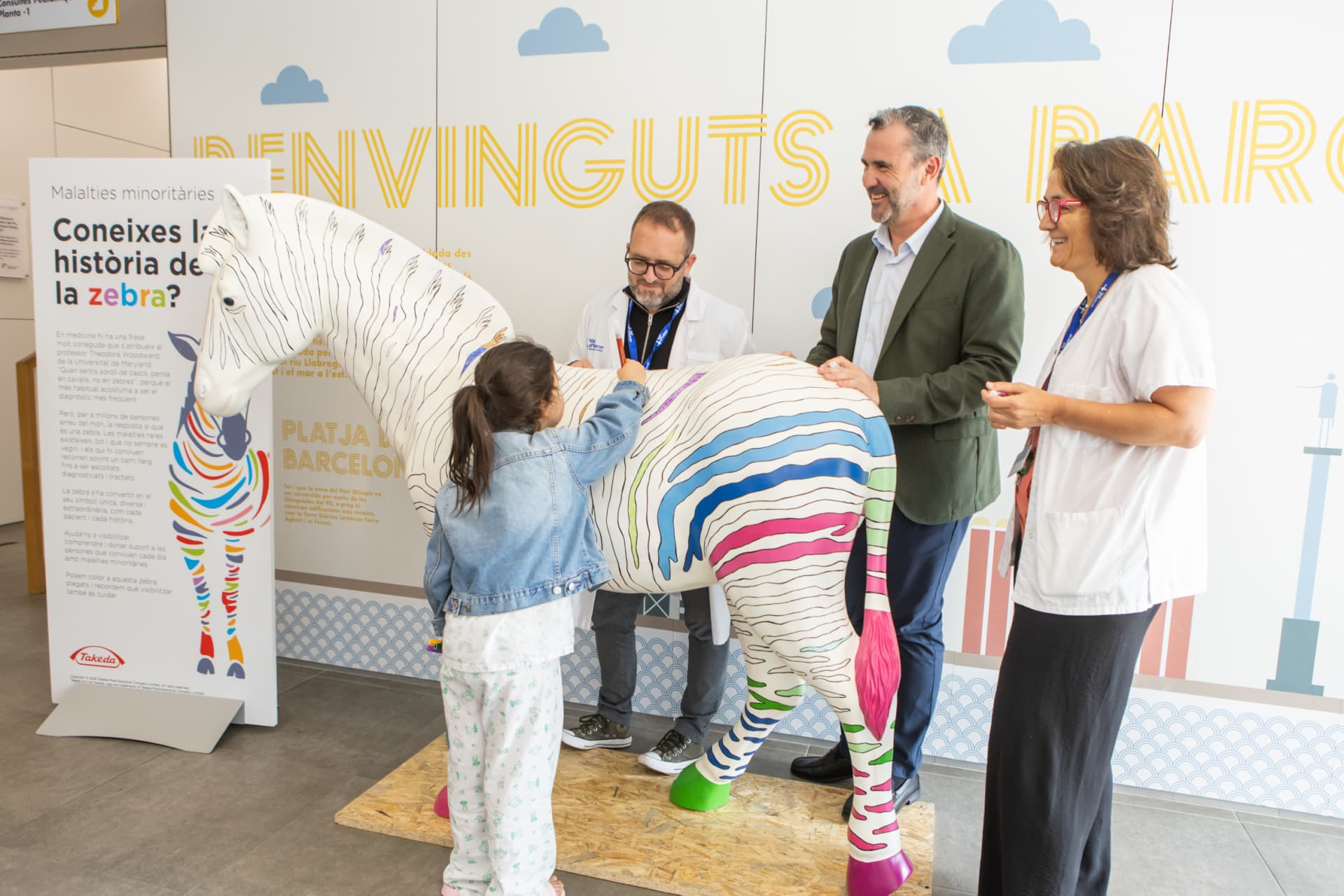 Photograph of a colorful zebra sculpture with rainbow stripes on legs, tail, and body, displayed indoors with four people interacting around it.