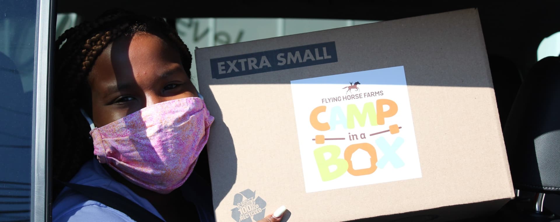 A child looking out from a window of a car wearing a mask and holding a package