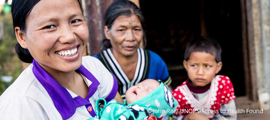Woman smiling carrying a baby. A child and a woman stand at the back