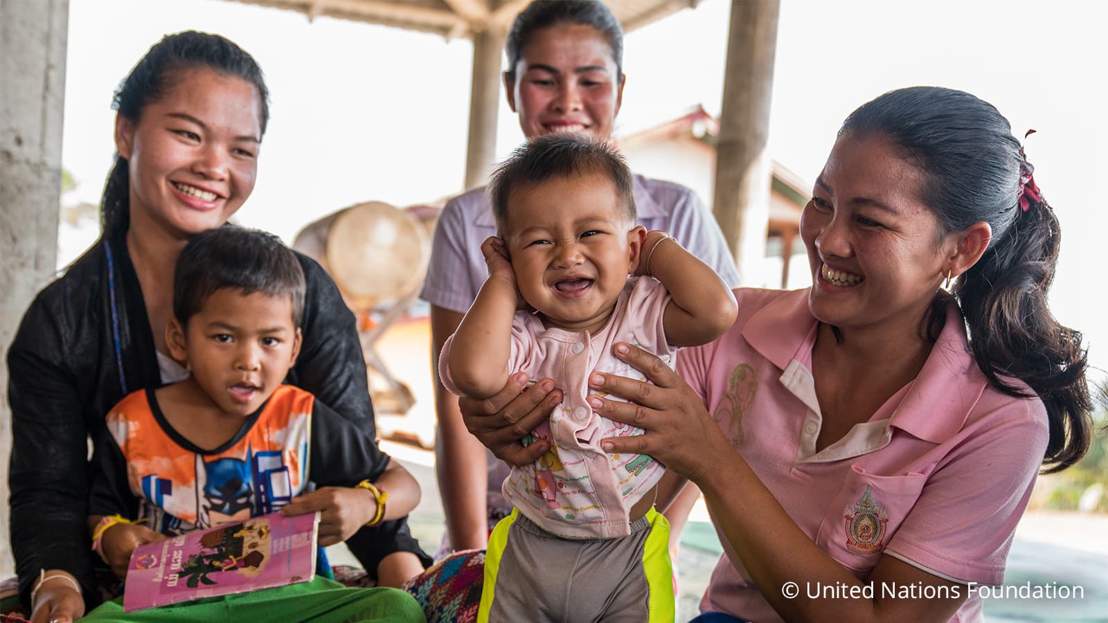 Woman holding boy. Another boy and two women at the background. All smiling.